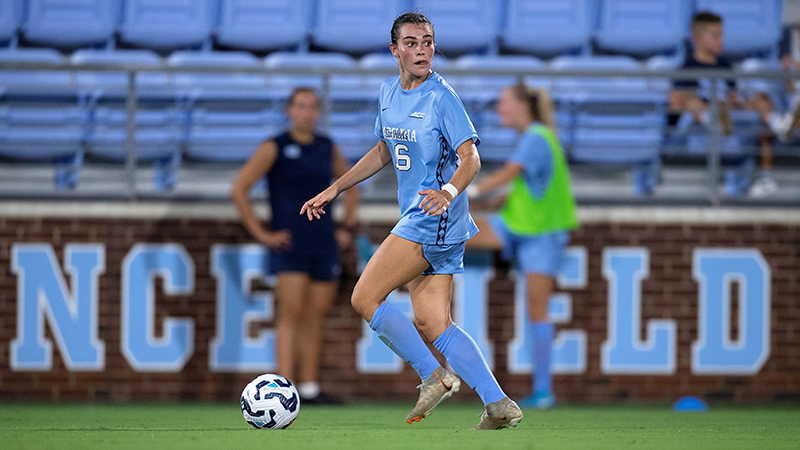 Female soccer player in light blue uniform looks over her shoulder ash she approaches the ball. other players and bleachers are blurred in the background.
