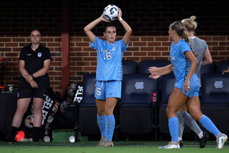 Female soccer player in light blue uniform holds ball over her head in lead up to throw, several other players approach from the right. Another, in a black uniform, watches from off field on the left.