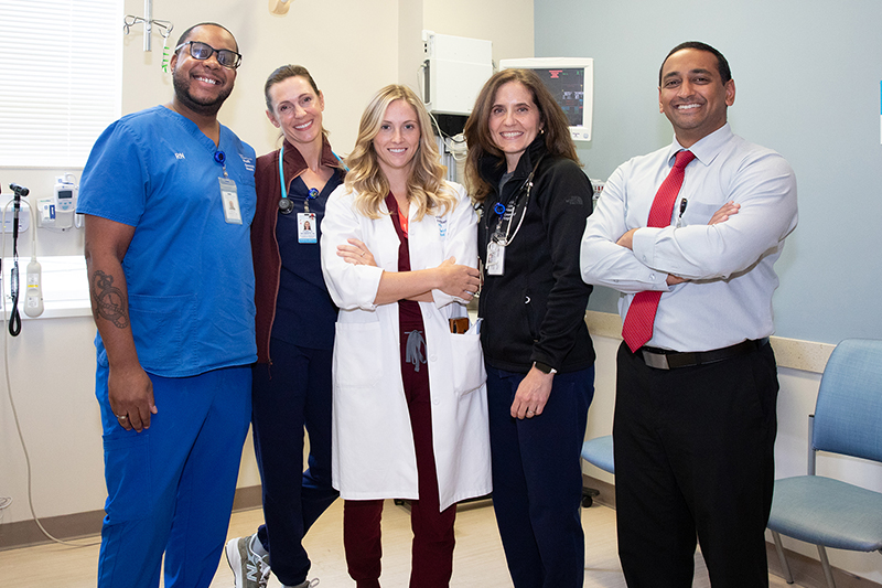 A group of four healthcare workers, in medical dress, pose in a treatment room; a fifth person, in business dress, stands to the right.