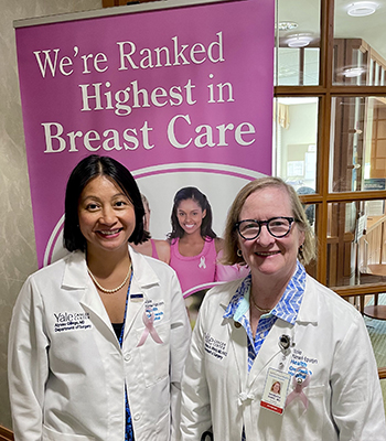 Two physicians in white clinical coats and wearing hospital id badges stand in front of a pink poster with the words We're Ranked Highest in Breast Care.