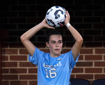 Woman in blue soccer uniform holds soccer ball over head in lead up to throw. Behind her is a brick facade.